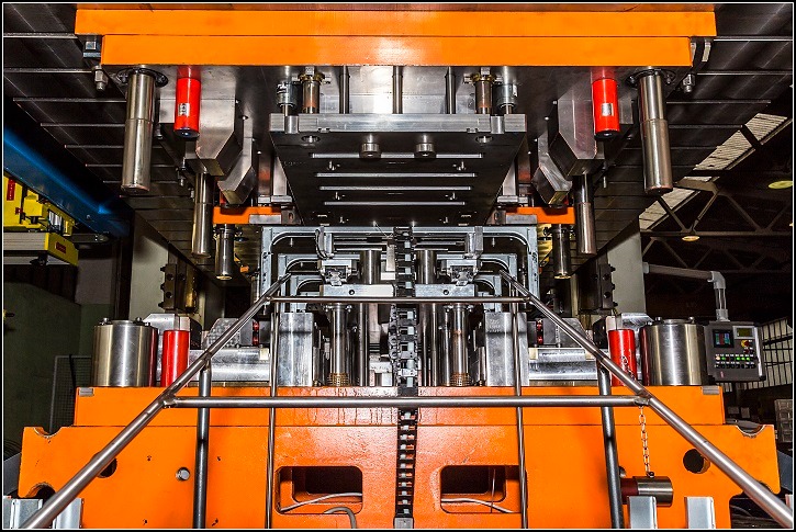 Widok z dołu na dużą, pomarańczowo-szarą prasę przemysłową z widocznymi elementami mocującymi, cylindrami hydraulicznymi i przewodami. Na drugim planie widoczny panel sterowania i oświetlenie...