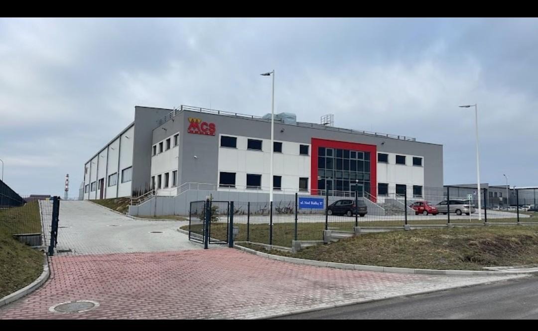 Budynek firmy MCS z logo na elewacji, widok z zewnątrz, z ogrodzeniem i wjazdem z kostki brukowej. Tabliczka z adresem 'ul. Nad Ralką 17'.