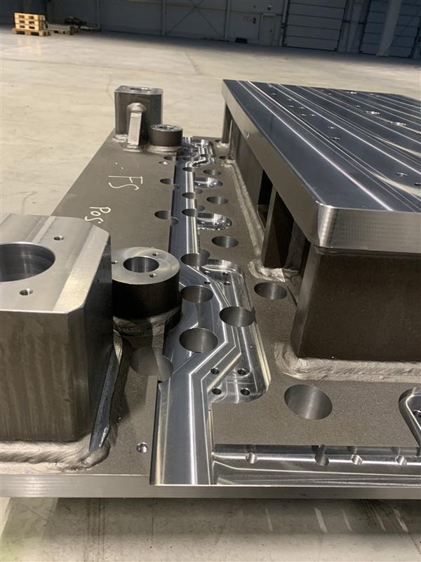 Precyzyjnie obrobiony, duży metalowy element z licznymi otworami i frezowaniami, widoczne ślady po obróbce skrawaniem, spawane elementy, napis 'Pos F5' na powierzchni, surowe oświetlenie hali.