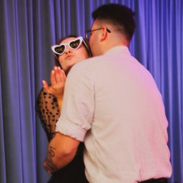 Monika Litwińska - Para w objęciach, kobieta w okularach w kształcie serc z napisem 'Cheers' posyła całusa, tło stanowią fioletowe zasłony.