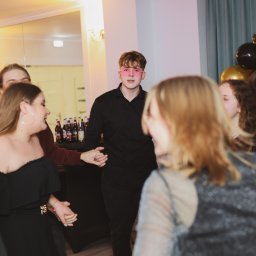 Monika Litwińska - Młody mężczyzna w czarnej koszuli i różowych okularach w kształcie serc prowadzi taniec z grupą uśmiechniętych osób na tle dekoracji z czarno-złotych balonów i butelek na stole.