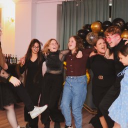 Monika Litwińska - Grupa rozbawionych nastolatków, w różnym wieku i ubraniach, tańczy i śmieje się na tle dekoracji z czarno-złotych balonów podczas imprezy urodzinowej.