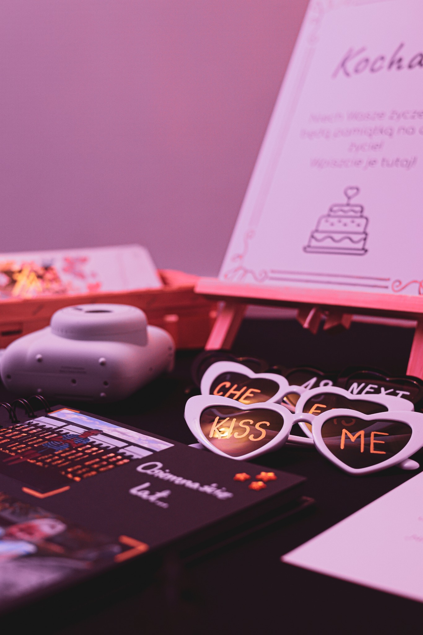 Dekoracje imprezowe w odcieniach różu: tablica z życzeniami z rysunkiem tortu, okulary w kształcie serc z napisem 'Kiss me', album ze zdjęciami z uroczystości.