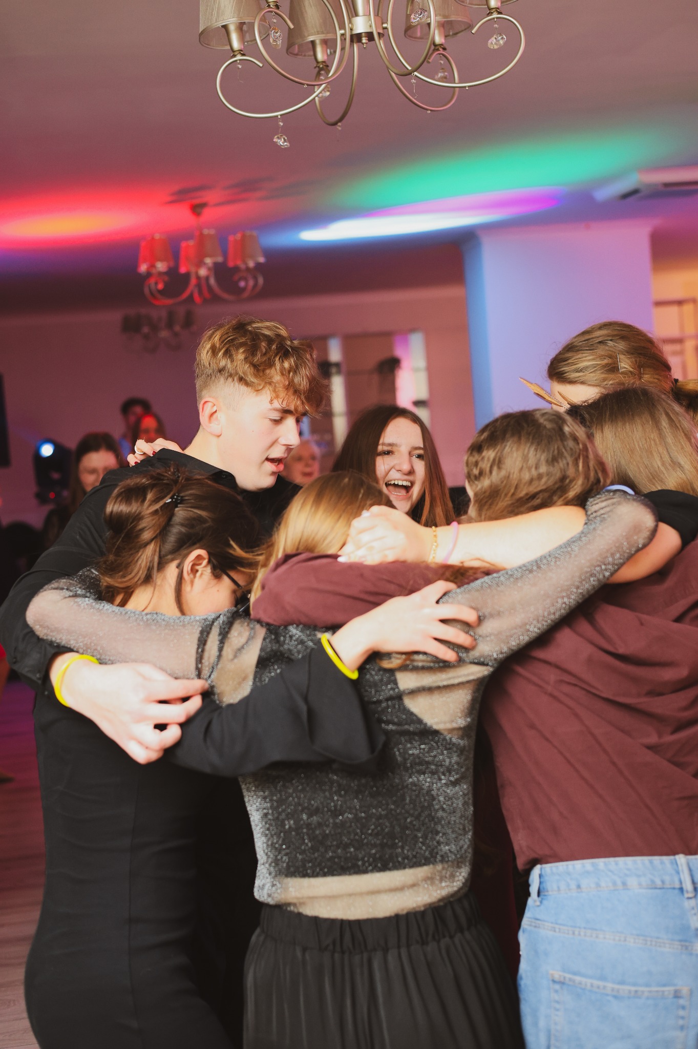 Grupa młodych ludzi w radosnym uścisku na tle sali z kolorowym oświetleniem, widoczne detale ubioru i fryzur.