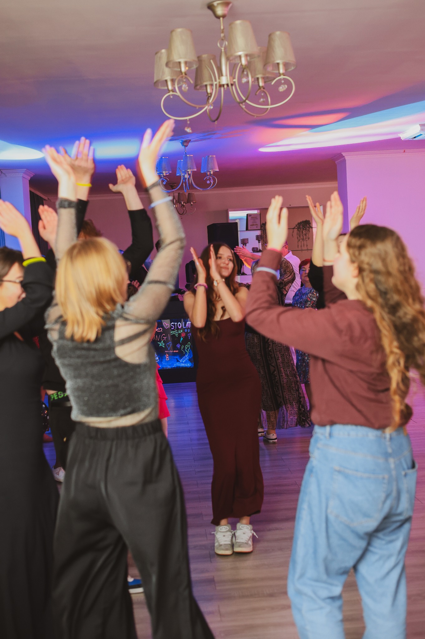 Grupa ludzi, w różnym wieku i ubraniach, z podniesionymi rękami, świętuje wydarzenie w sali oświetlonej kolorowymi światłami, z widocznym napisem 'Sto lat' na dekoracji.