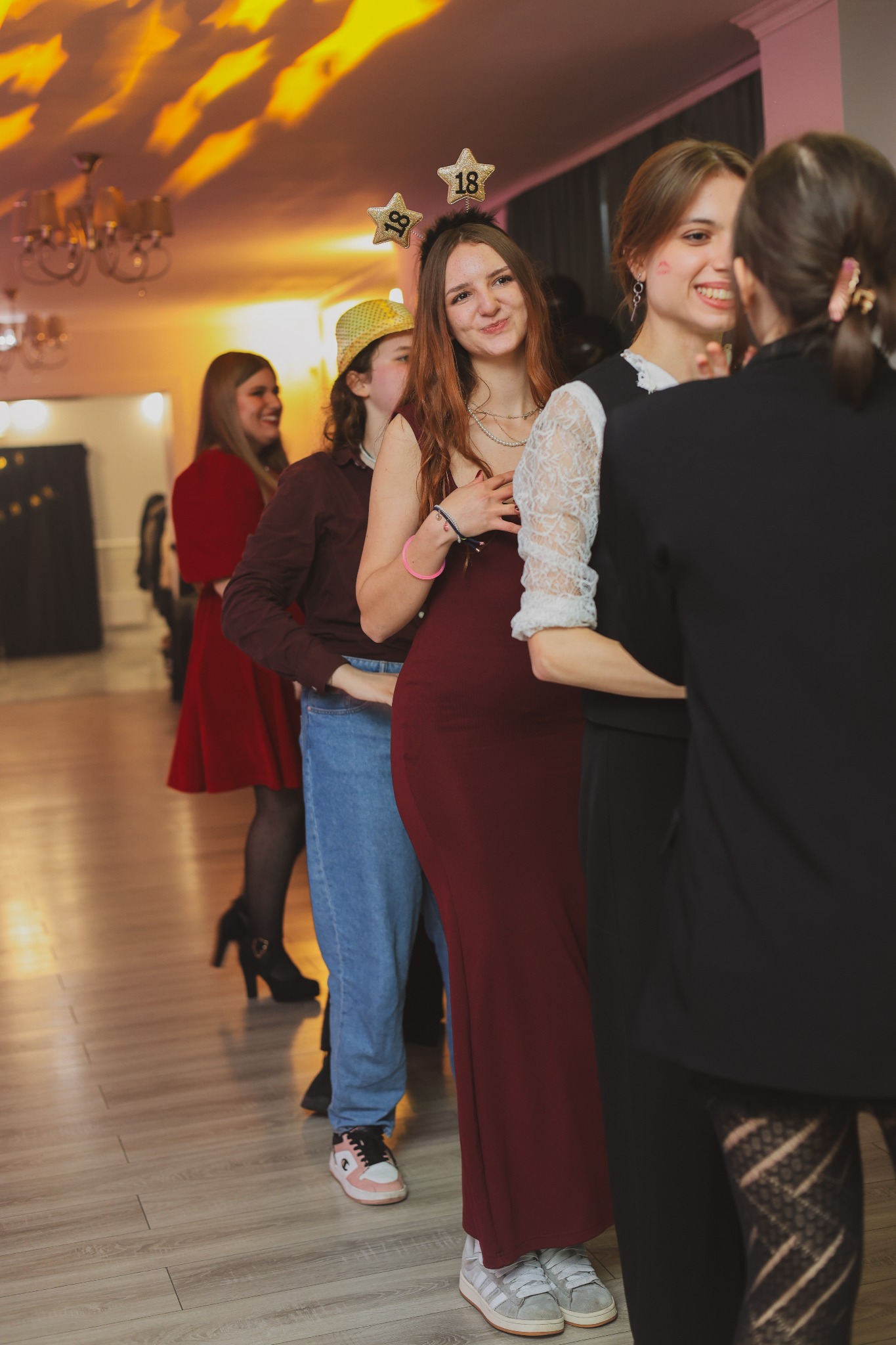 Grupa młodych kobiet w strojach imprezowych ustawiona w rzędzie, prawdopodobnie podczas świętowania osiemnastych urodzin, widoczna dekoracja w postaci gwiazdek z numerem 18 na głowie jednej...