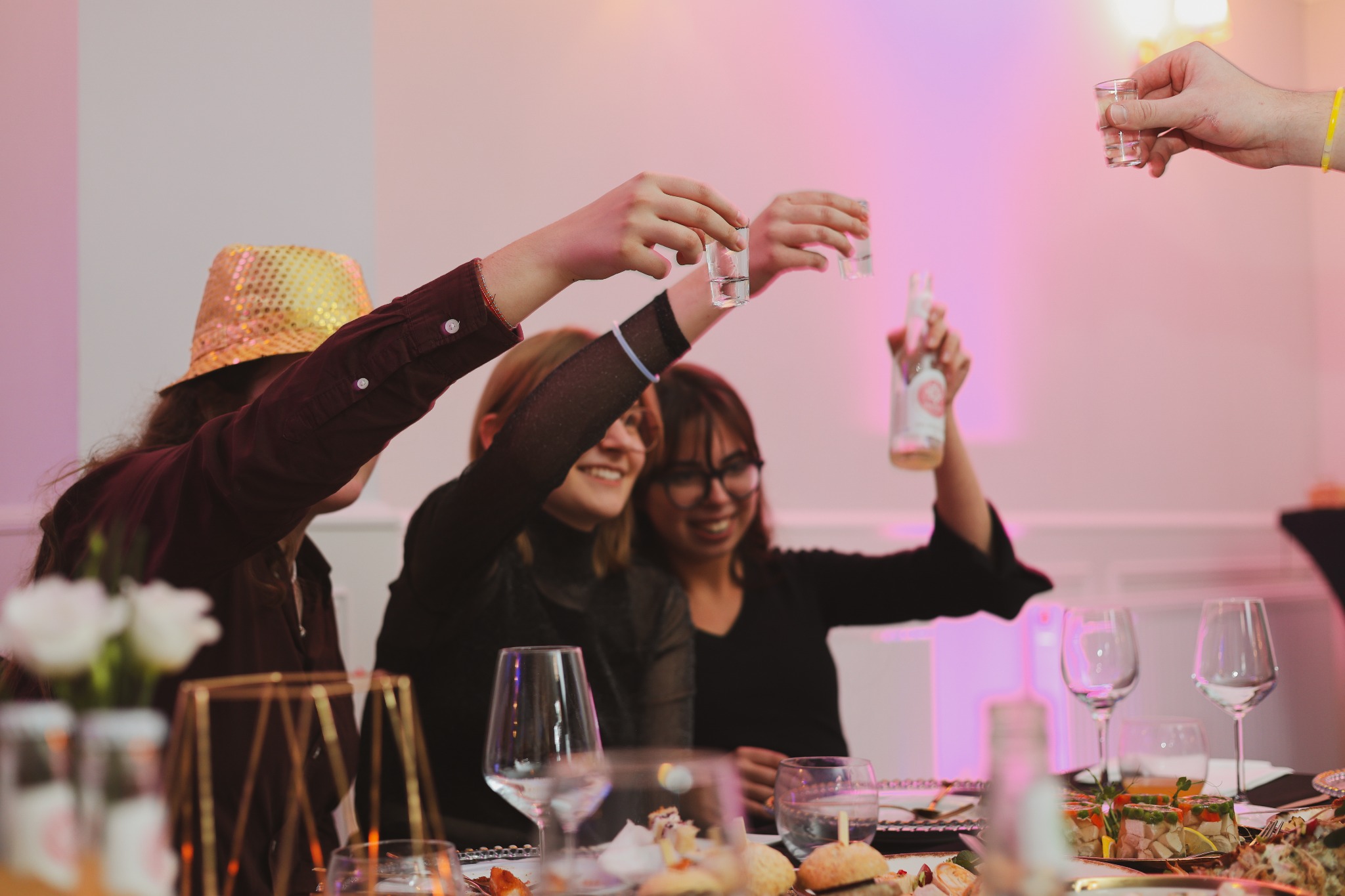 Grupa osób wznosi toast małymi kieliszkami nad bogato zastawionym stołem podczas imprezy, jedna osoba w złotym kapeluszu.