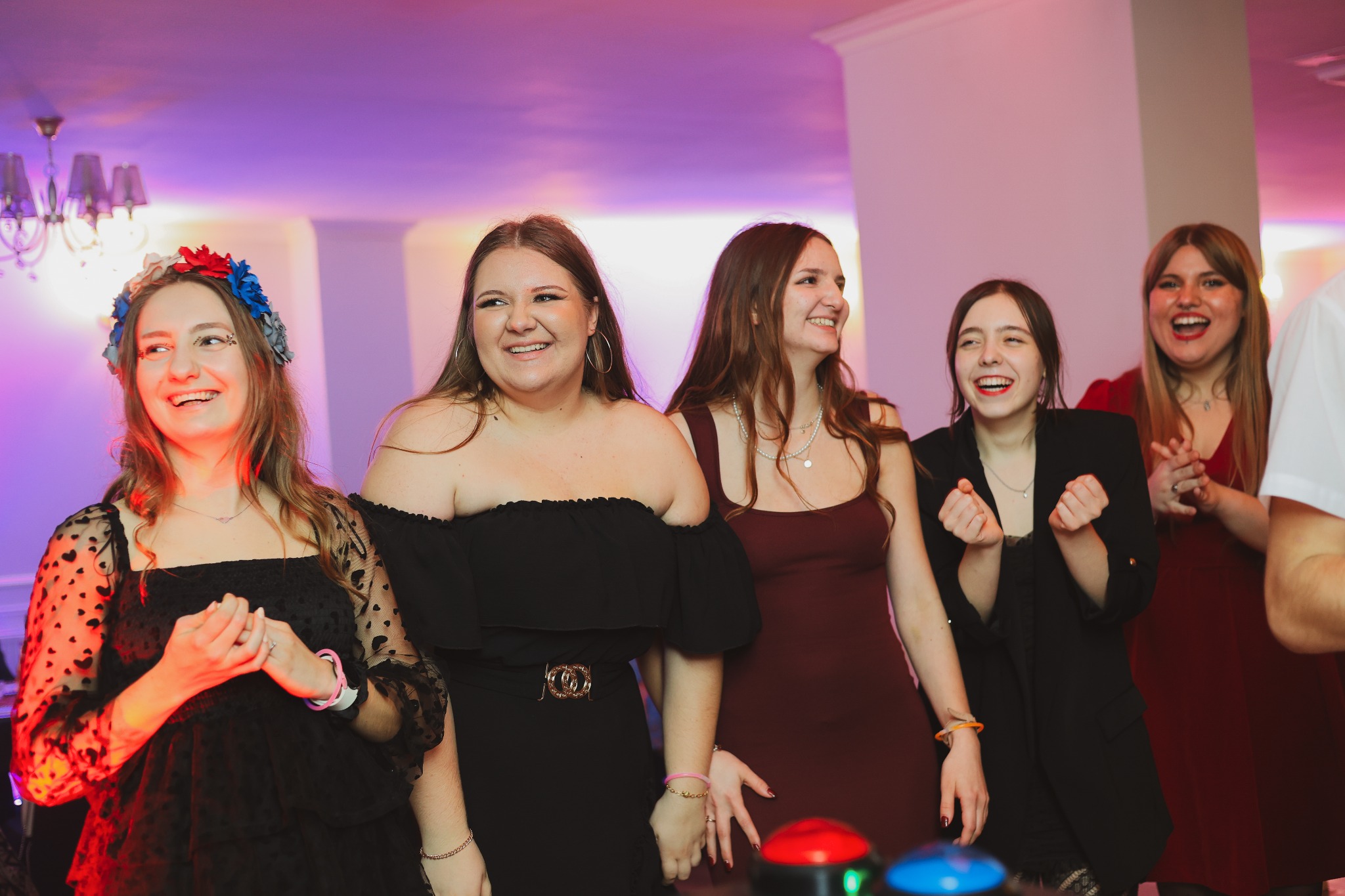 Grupa pięciu uśmiechniętych młodych kobiet w wieczorowych strojach, jedna z wiankiem na głowie, w jasnym pomieszczeniu z kolorowym oświetleniem i widocznymi przyciskami do gry.