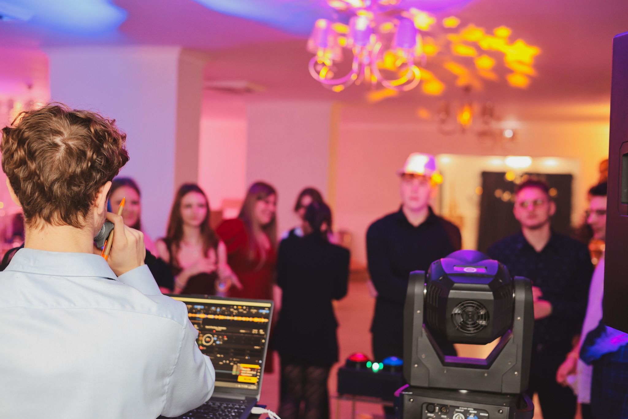 DJ z mikrofonem obsługujący laptopa na tle bawiących się gości i oświetlenia eventowego.