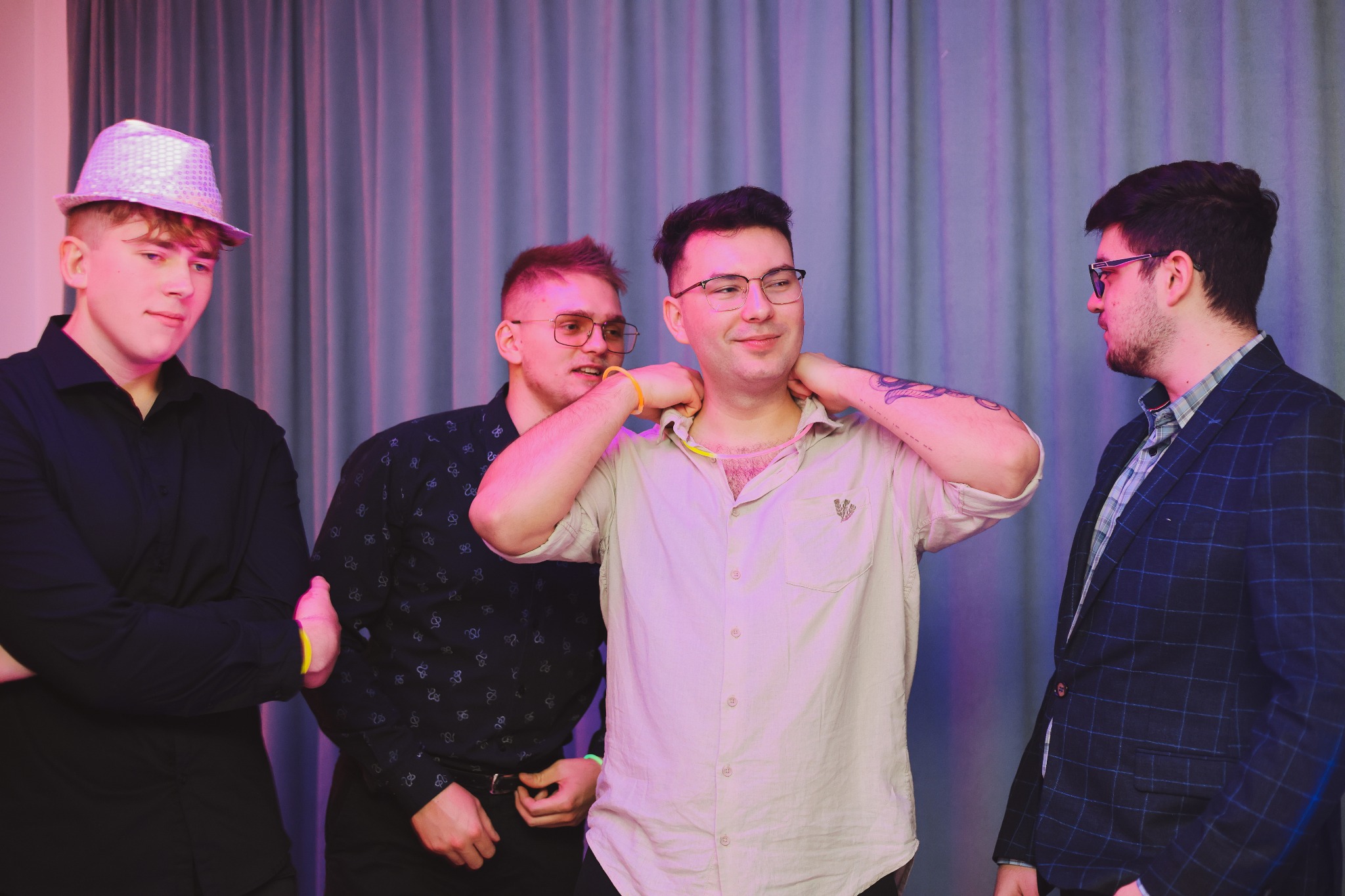 Grupa czterech mężczyzn w różnym wieku i strojach pozuje na tle fioletowej zasłony, jeden z nich w błyszczącym kapeluszu, inny w rozpiętej koszuli z tatuażem na ramieniu.