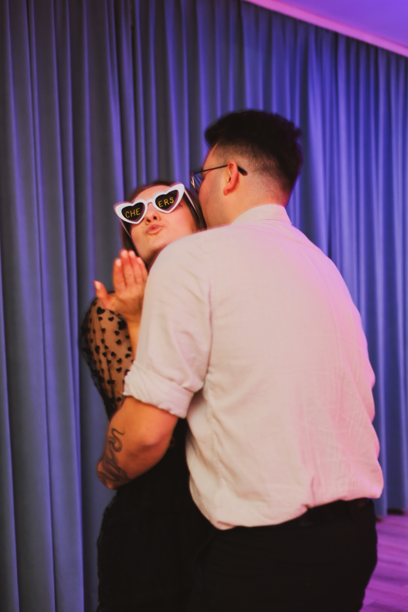 Para w objęciach, kobieta w okularach w kształcie serc z napisem 'Cheers' posyła całusa, tło stanowią fioletowe zasłony.