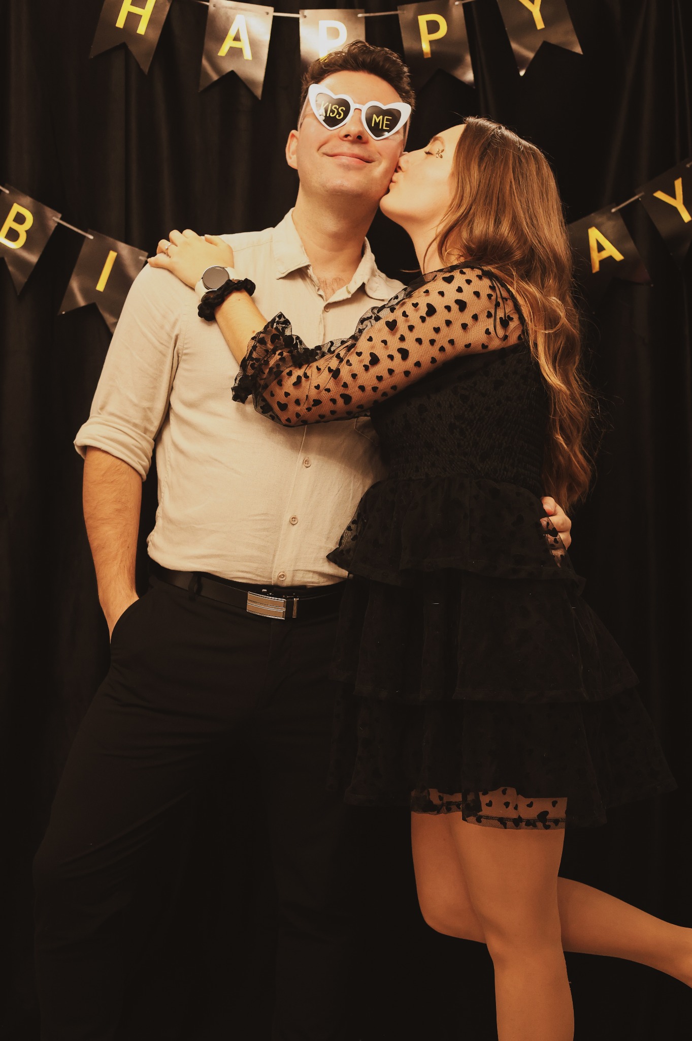 Kobieta w czarnej sukience w serduszka całuje mężczyznę w okularach w kształcie serc z napisem 'Kiss Me' na tle czarnej kurtyny z napisem 'Happy Birthday'.