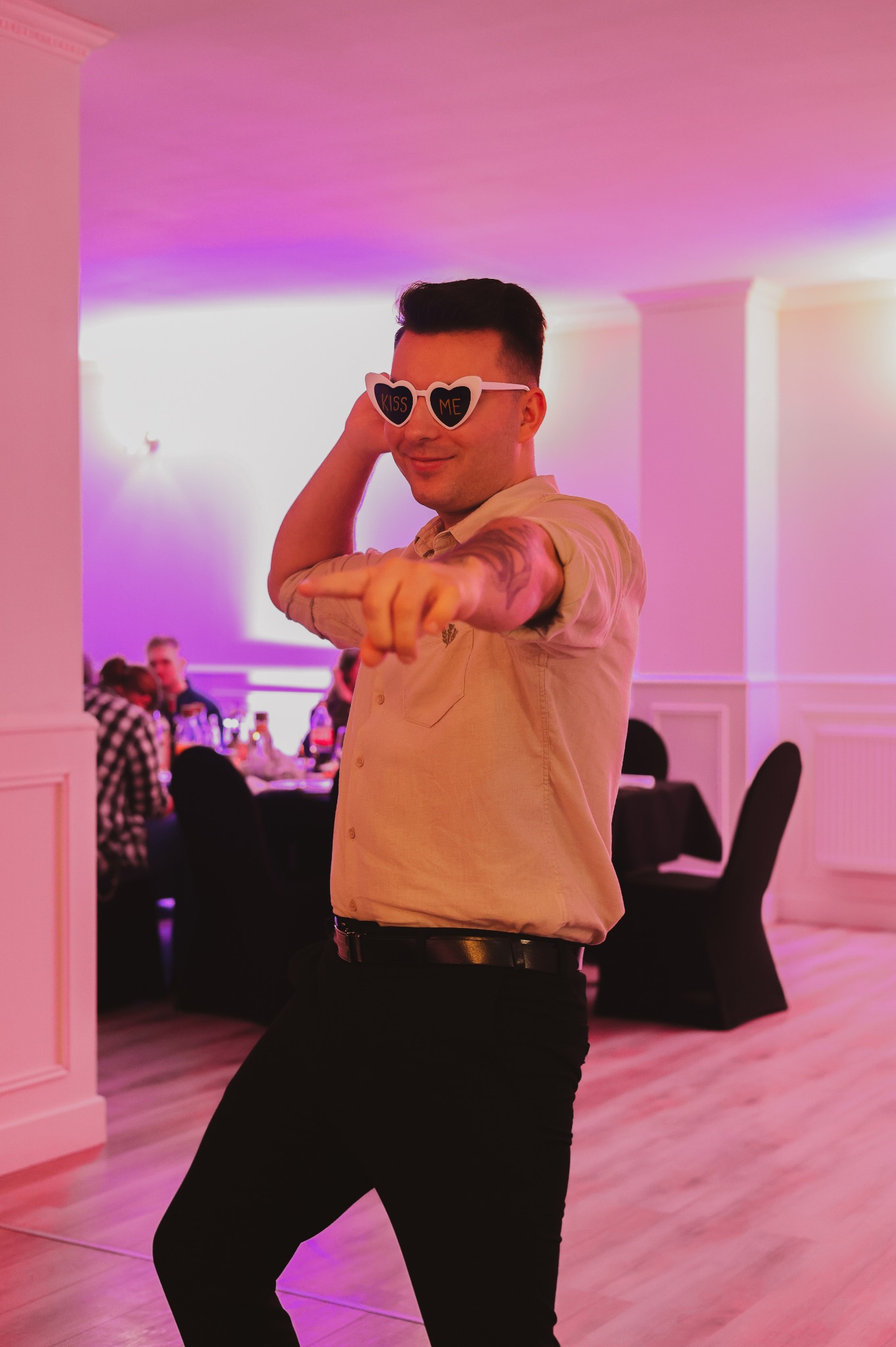 Mężczyzna w okularach w kształcie serc z napisem 'Kiss Me' tańczy na imprezie, wskazując palcem w stronę widza, w tle widać gości przy stole, oświetlenie w odcieniach różu.