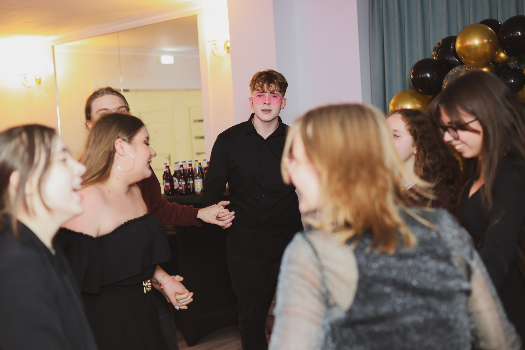 Młody mężczyzna w czarnej koszuli i różowych okularach w kształcie serc prowadzi taniec z grupą uśmiechniętych osób na tle dekoracji z czarno-złotych balonów i butelek na stole.