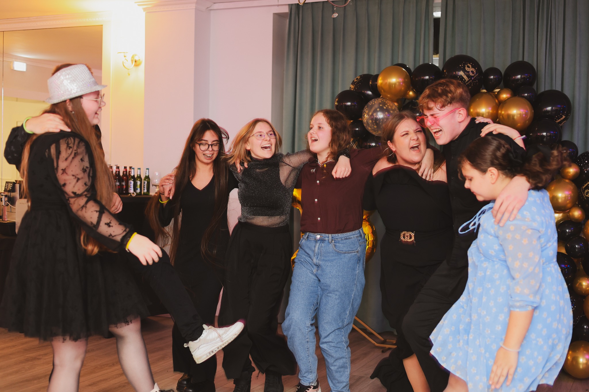 Grupa rozbawionych nastolatków, w różnym wieku i ubraniach, tańczy i śmieje się na tle dekoracji z czarno-złotych balonów podczas imprezy urodzinowej.