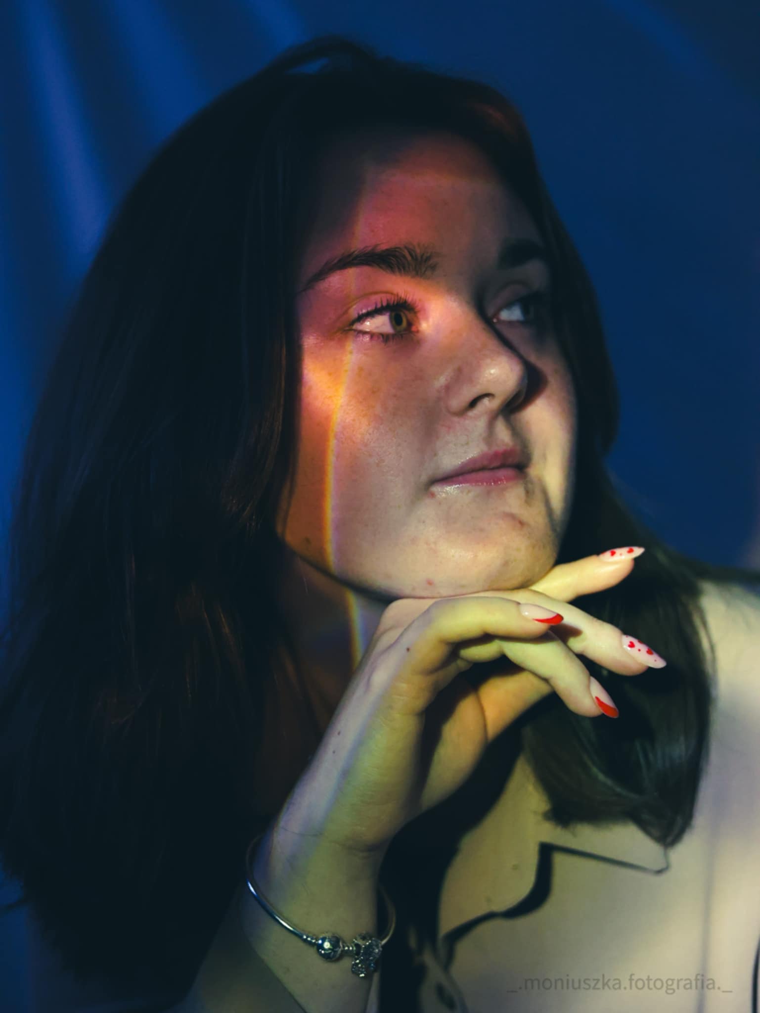 Portret kobiety z tęczowym światłem na twarzy, opierającej brodę na dłoni z czerwonymi paznokciami, na ciemnym niebieskim tle.