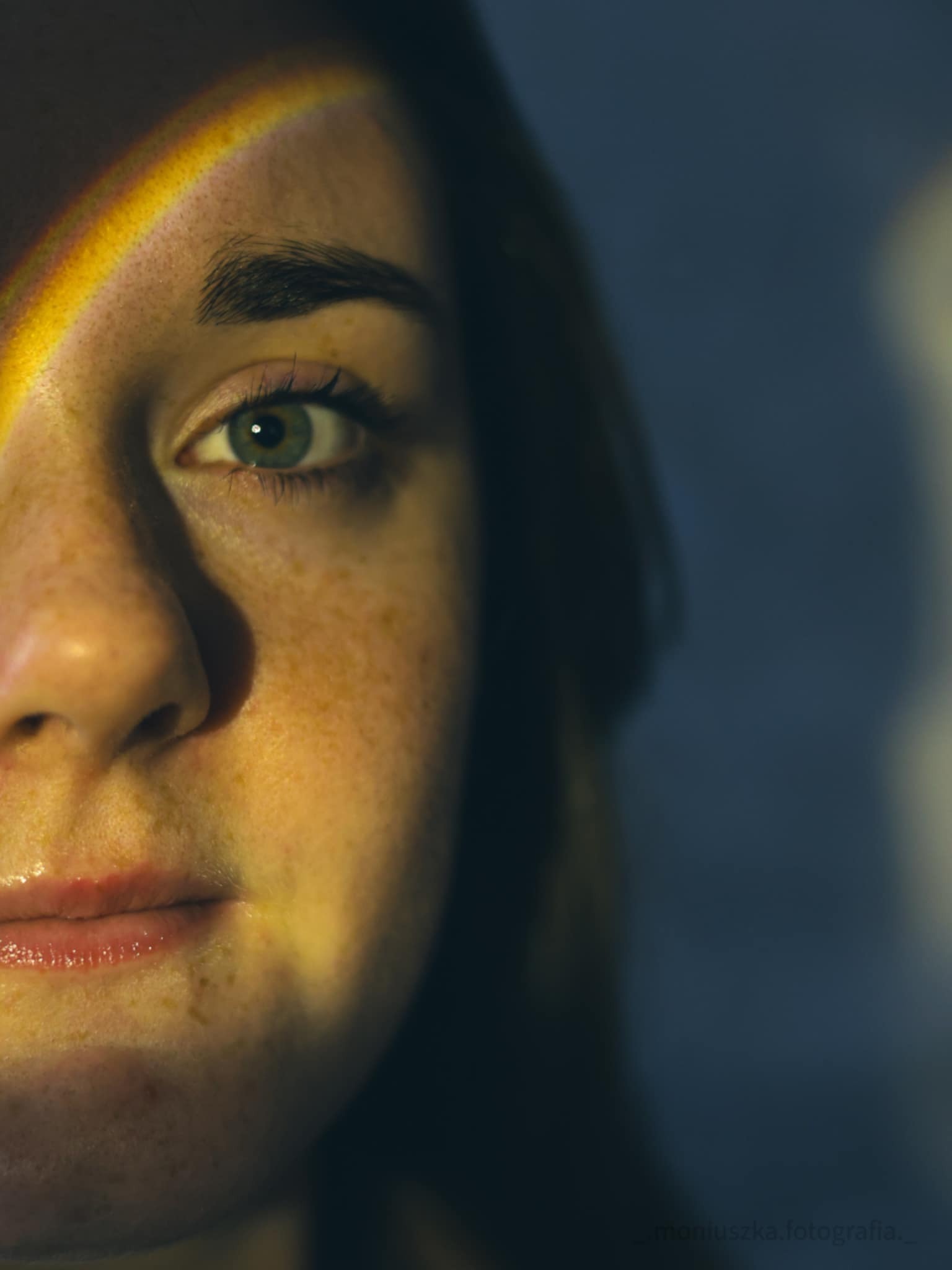 Portret zbliżeniowy kobiety z piegami, oświetlonej tęczowym światłem na twarzy, z zielonym okiem skierowanym wprost, na ciemnym tle.