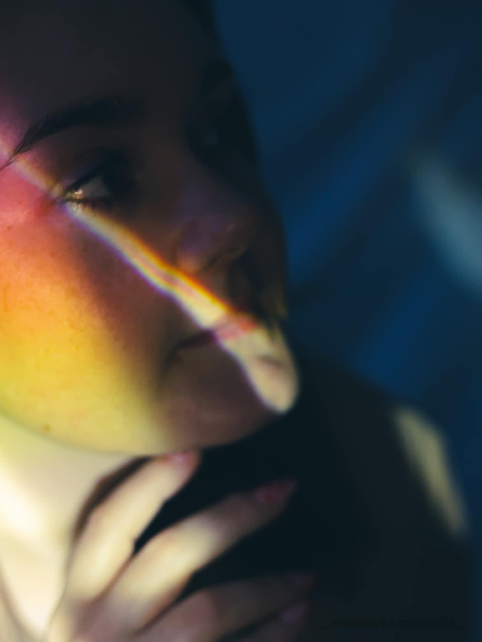 Portret kobiety z tęczowym światłem na twarzy, delikatna dłoń z różowym manicure w kadrze, ciemne tło.