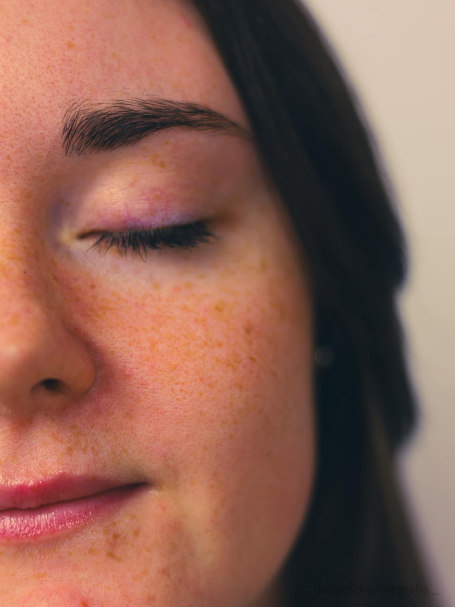 Portret fragmentu twarzy kobiety z zamkniętym okiem, widoczne piegi i delikatny makijaż oka w odcieniach fioletu.