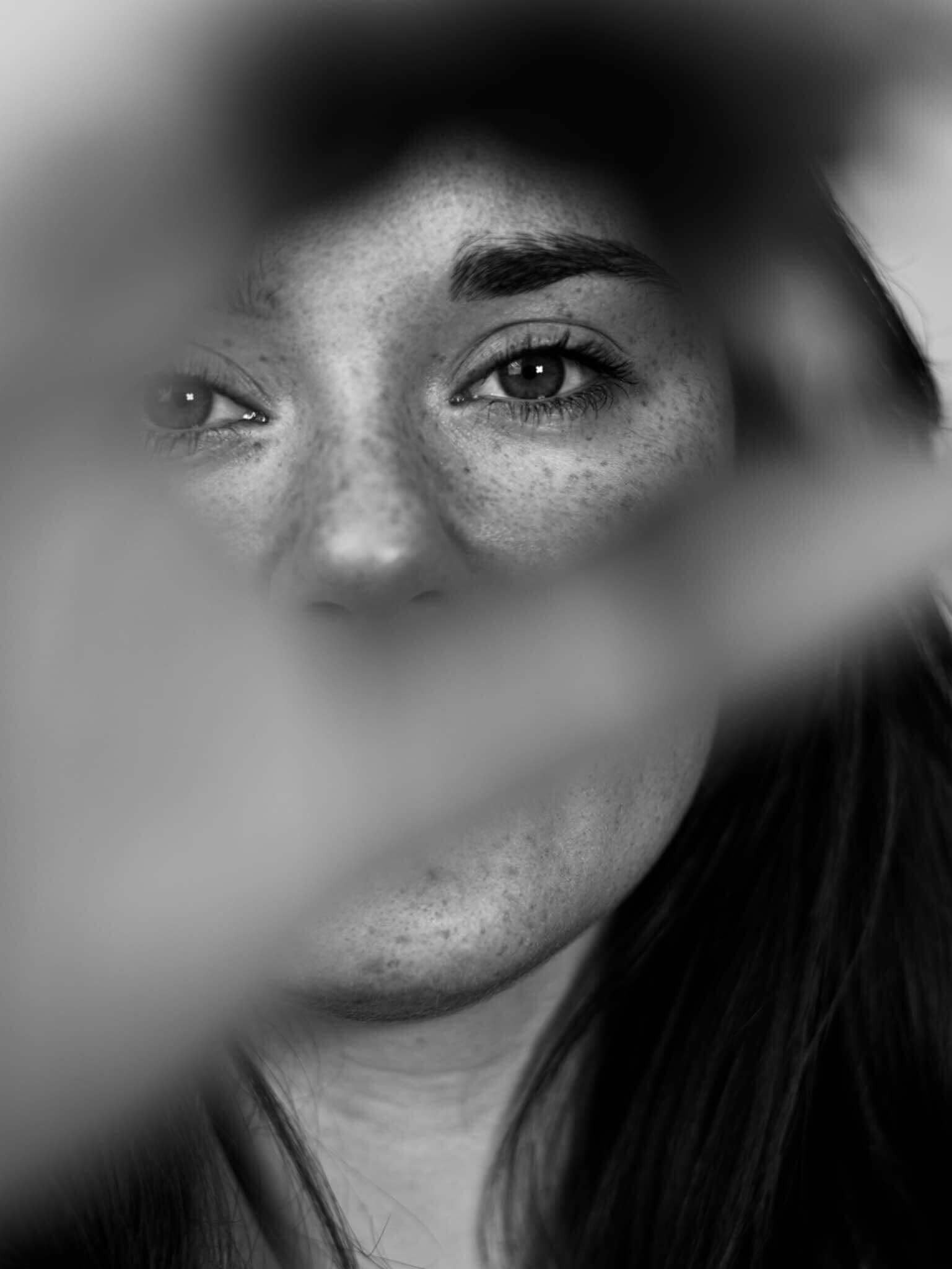Czarno-białe zbliżenie na twarz kobiety z piegami, częściowo zasłoniętą rozmytym elementem, skupiające się na oczach i brwiach.