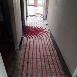 Mateusz Młynarski Technika Grzewcza - Różowe rurki ogrzewania podłogowego ułożone na folii z siatką w surowym wnętrzu, widok z perspektywy korytarza w kierunku przeszklonych drzwi.