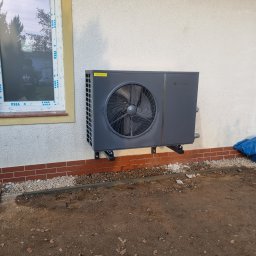 Mateusz Młynarski Technika Grzewcza - Szara pompa ciepła zamontowana na ścianie budynku z ceglaną podmurówką, widoczna z zewnątrz, z oknem w tle.