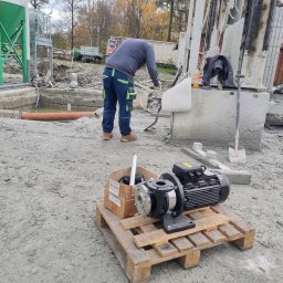 Mateusz Młynarski Technika Grzewcza - Montaż pompy ciepła: pracownik w ubraniu roboczym przy wiertnicy, pompa i akcesoria na palecie, zbiornik i instalacja w tle.