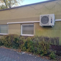 Mateusz Młynarski Technika Grzewcza - Zewnętrzna jednostka klimatyzacji marki AUX zamontowana na elewacji budynku w Gryfowie Śląskim, widoczna z chodnika, z krzewami u podstawy ściany.