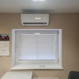 Biała klimatyzacja na ścianie nad oknem z żaluzjami w pomieszczeniu biurowym, widoczny kalendarz i fragment biurka.