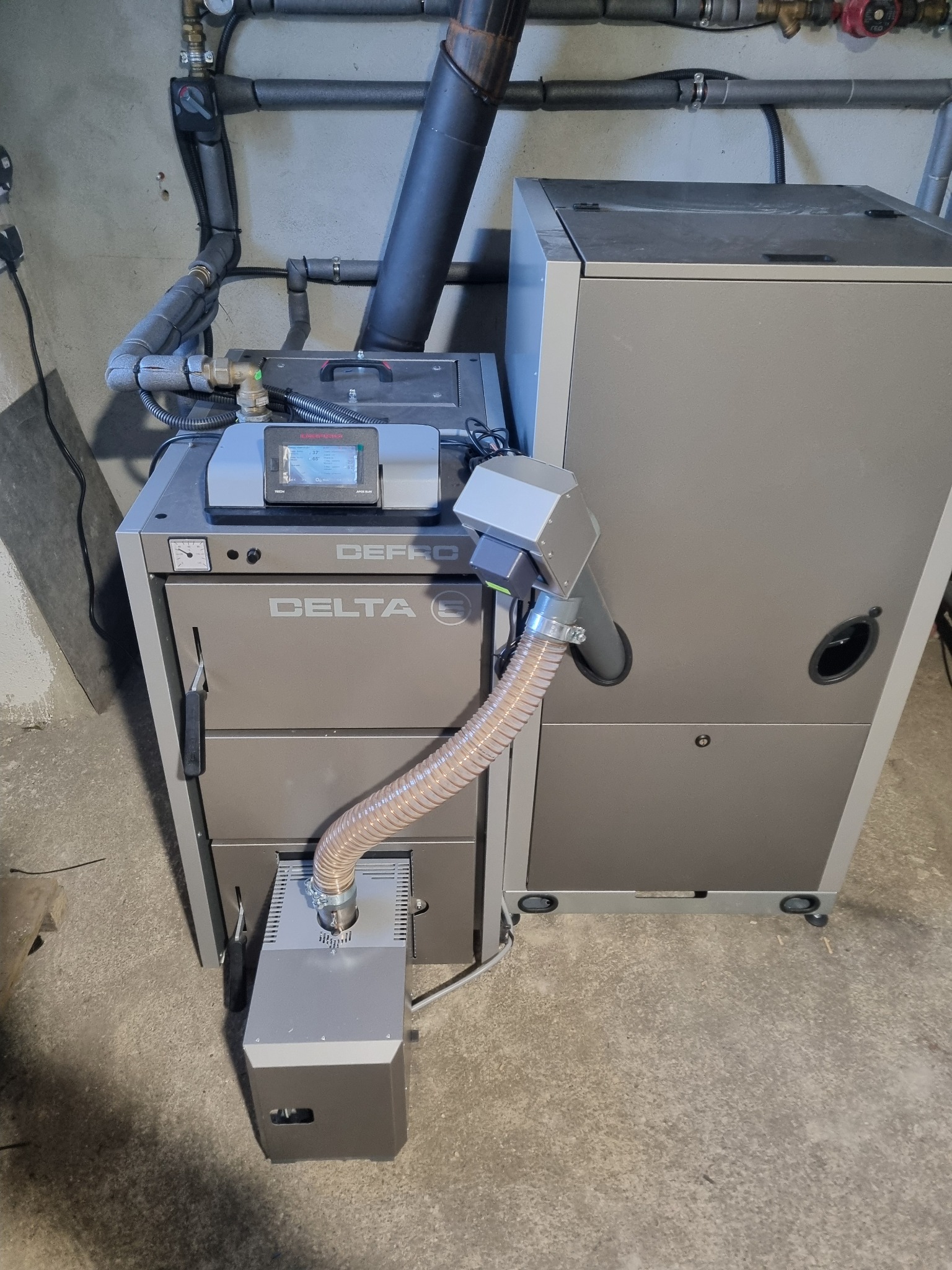 Szary piec CO marki Defro Delta z podajnikiem, rura spalinowa i instalacja hydrauliczna w tle.