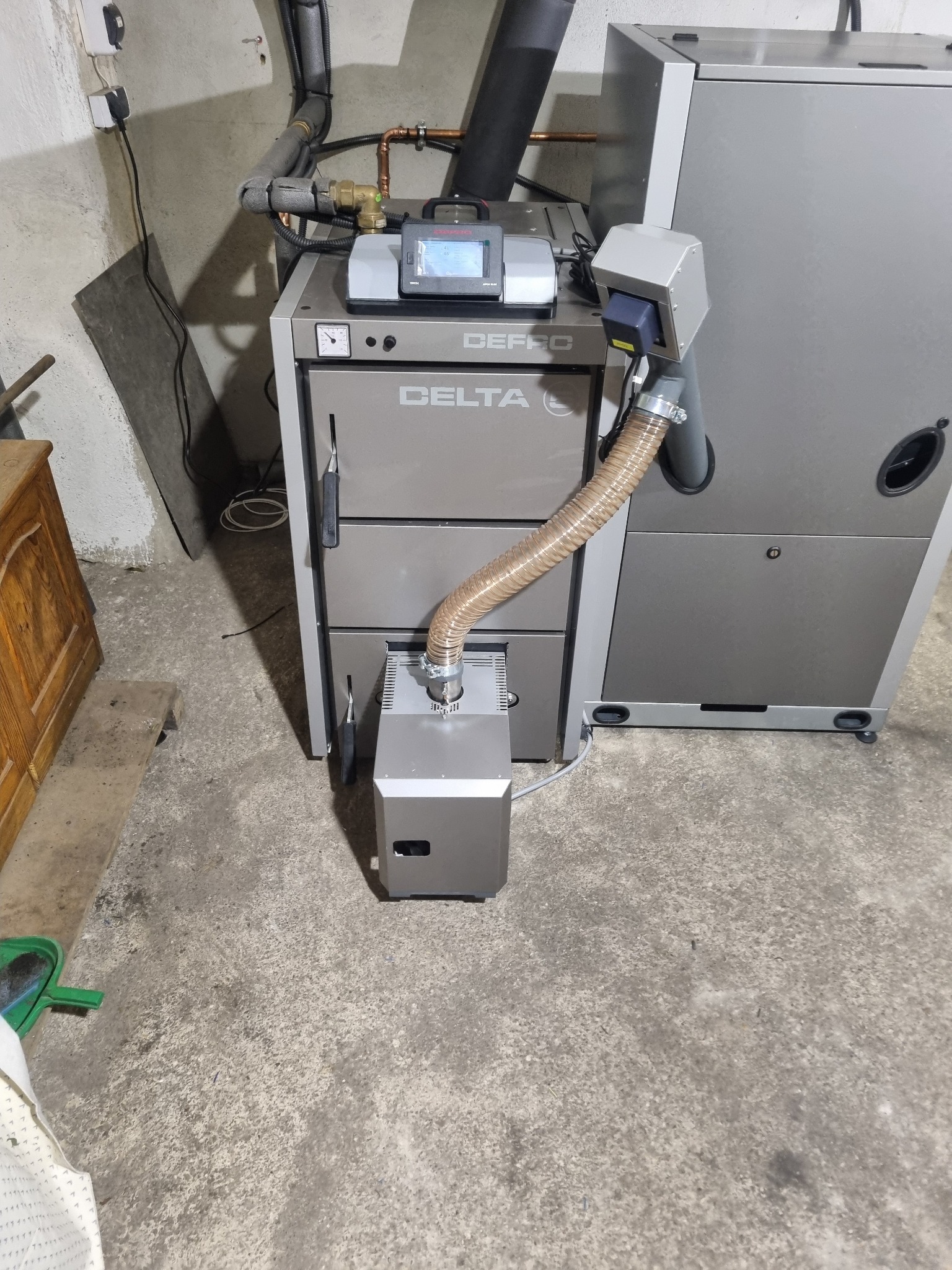 Szary piec CO marki Defro z podajnikiem ślimakowym, sterownikiem i przewodami w kotłowni z betonową posadzką.