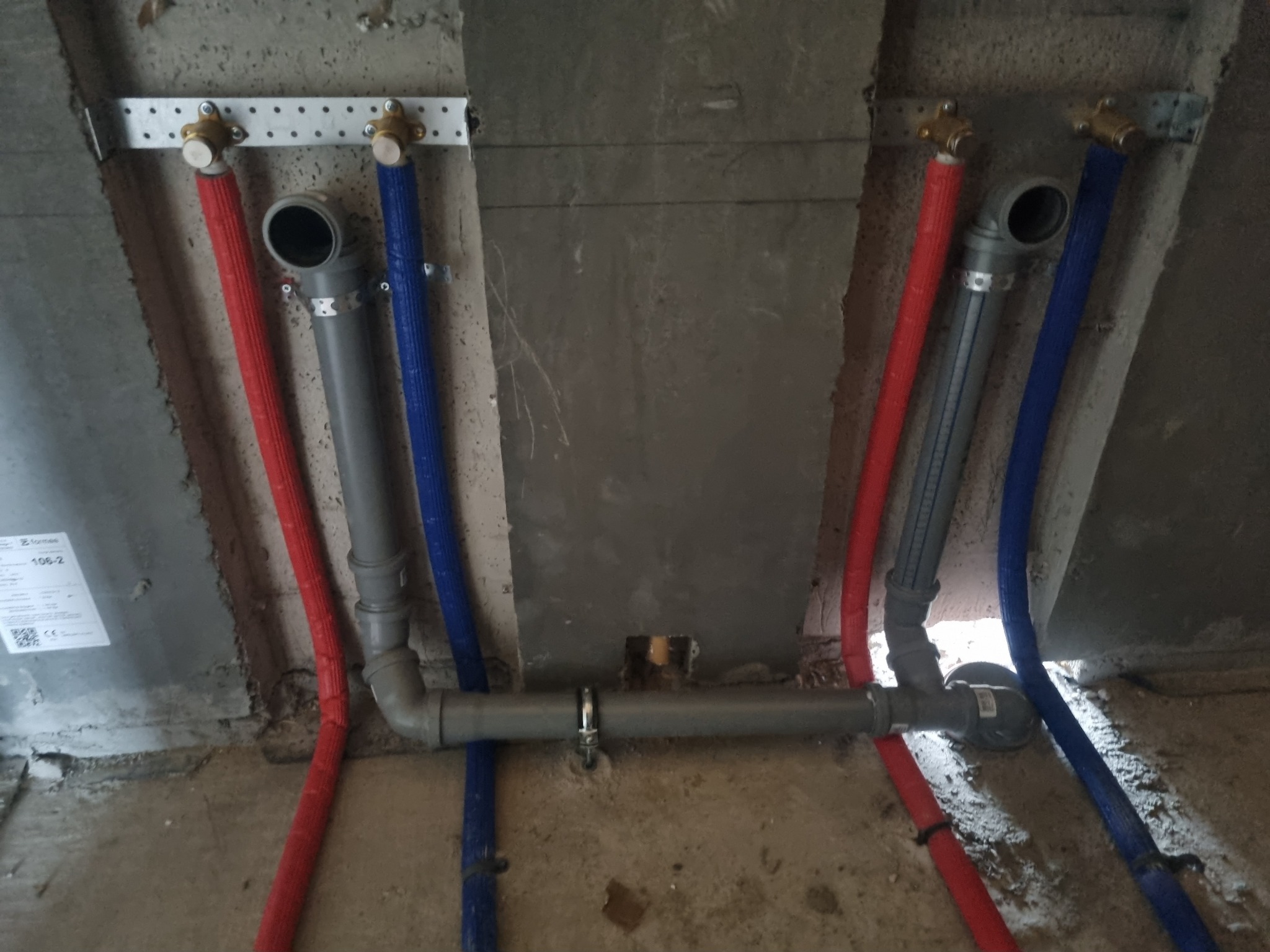 Szare rury kanalizacyjne połączone z czerwonymi i niebieskimi rurami w izolacji, zamontowane na surowym, betonowym tle ściany w trakcie prac instalacyjnych.