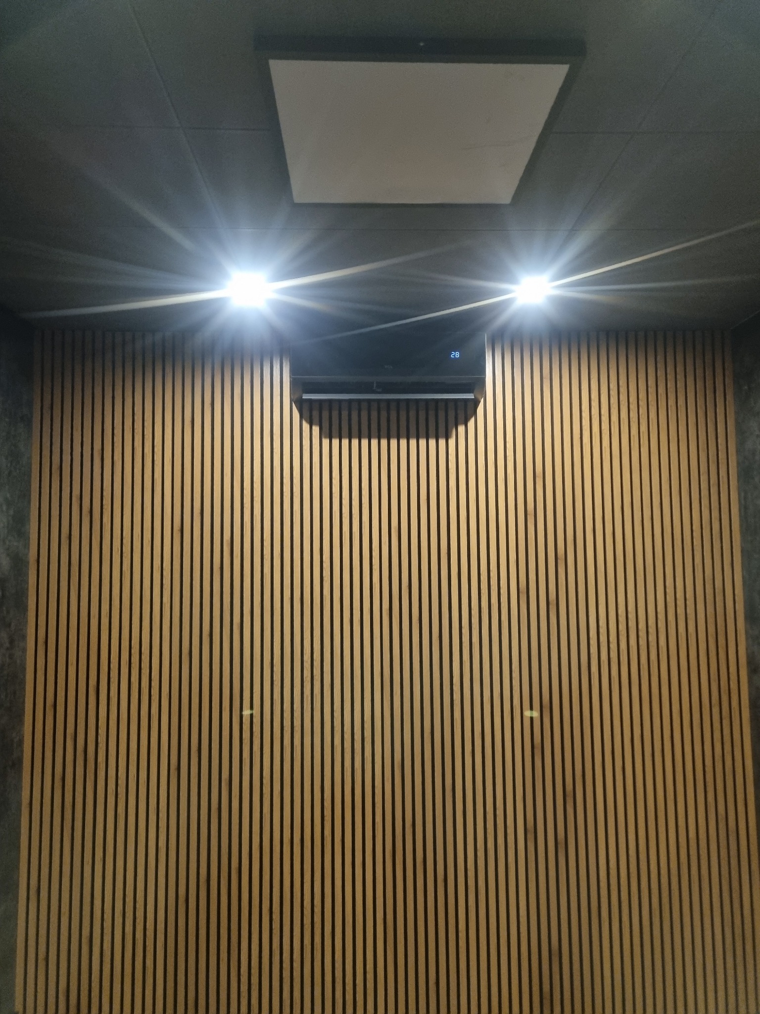 Klimatyzator ścienny włączony, wyświetlający temperaturę 28 stopni, zamontowany na ścianie z drewnianych listewek, oświetlony dwoma halogenami