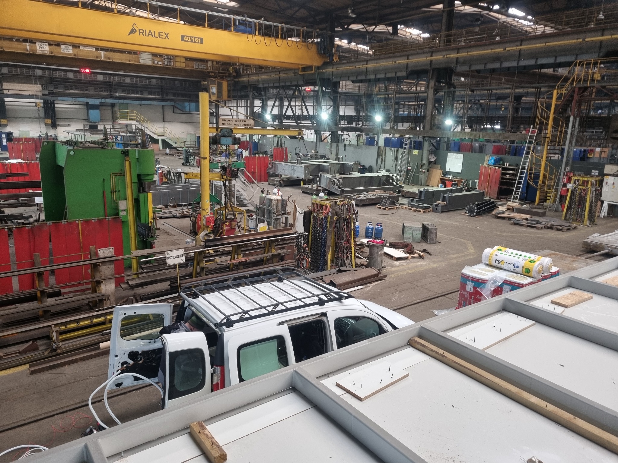 Wnętrze hali produkcyjnej z suwnicą, widok z góry na biały samochód dostawczy z otwartymi drzwiami, materiały izolacyjne i konstrukcję szkieletową w pierwszym planie.