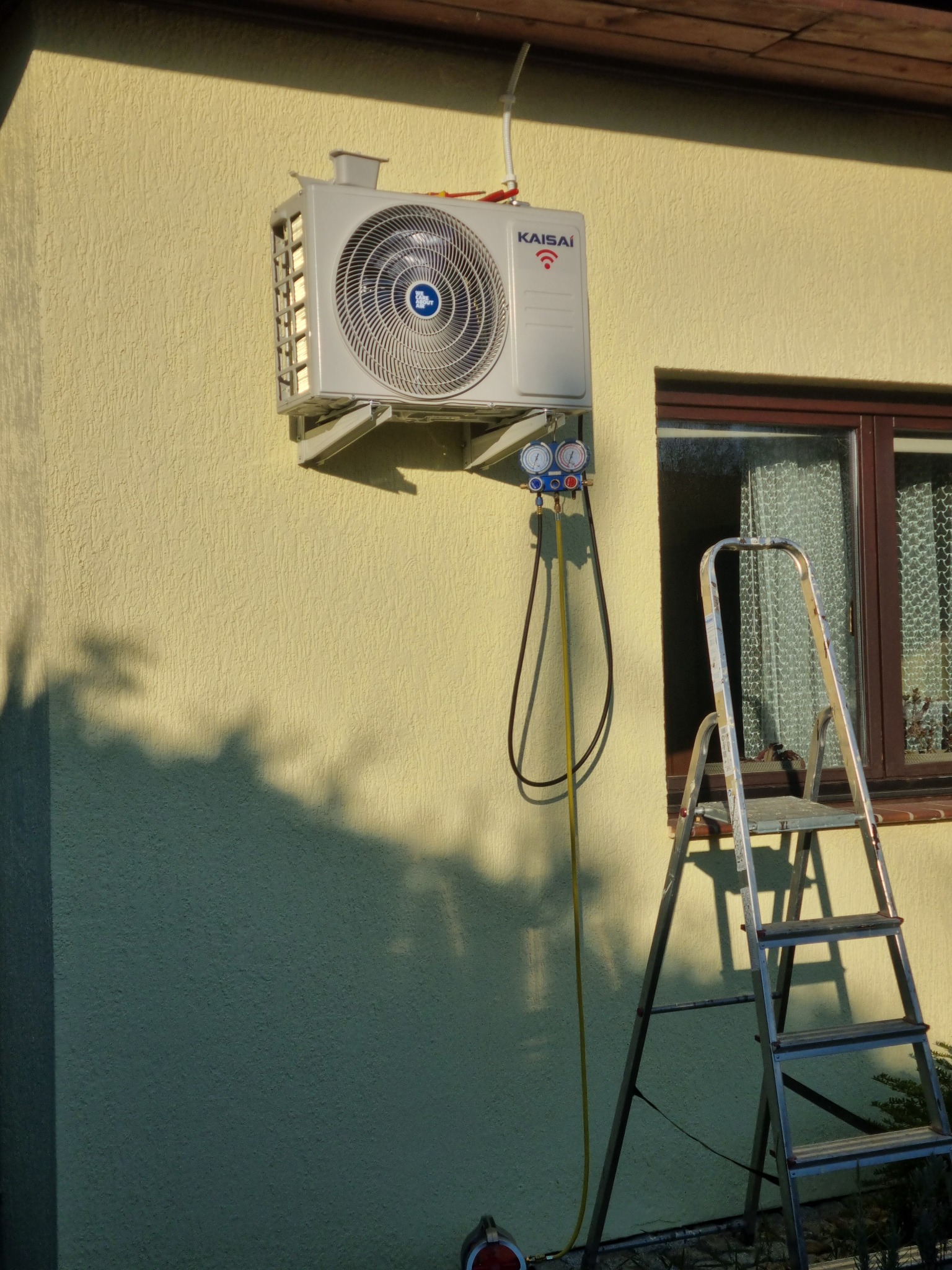 Zewnętrzna jednostka klimatyzacji Kaisai zamontowana na elewacji budynku, podłączona do manometrów i węży serwisowych, obok drabina aluminiowa.