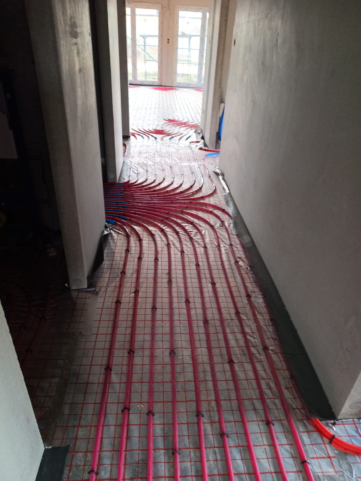 Różowe rurki ogrzewania podłogowego ułożone na folii z siatką w surowym wnętrzu, widok z perspektywy korytarza w kierunku przeszklonych drzwi.