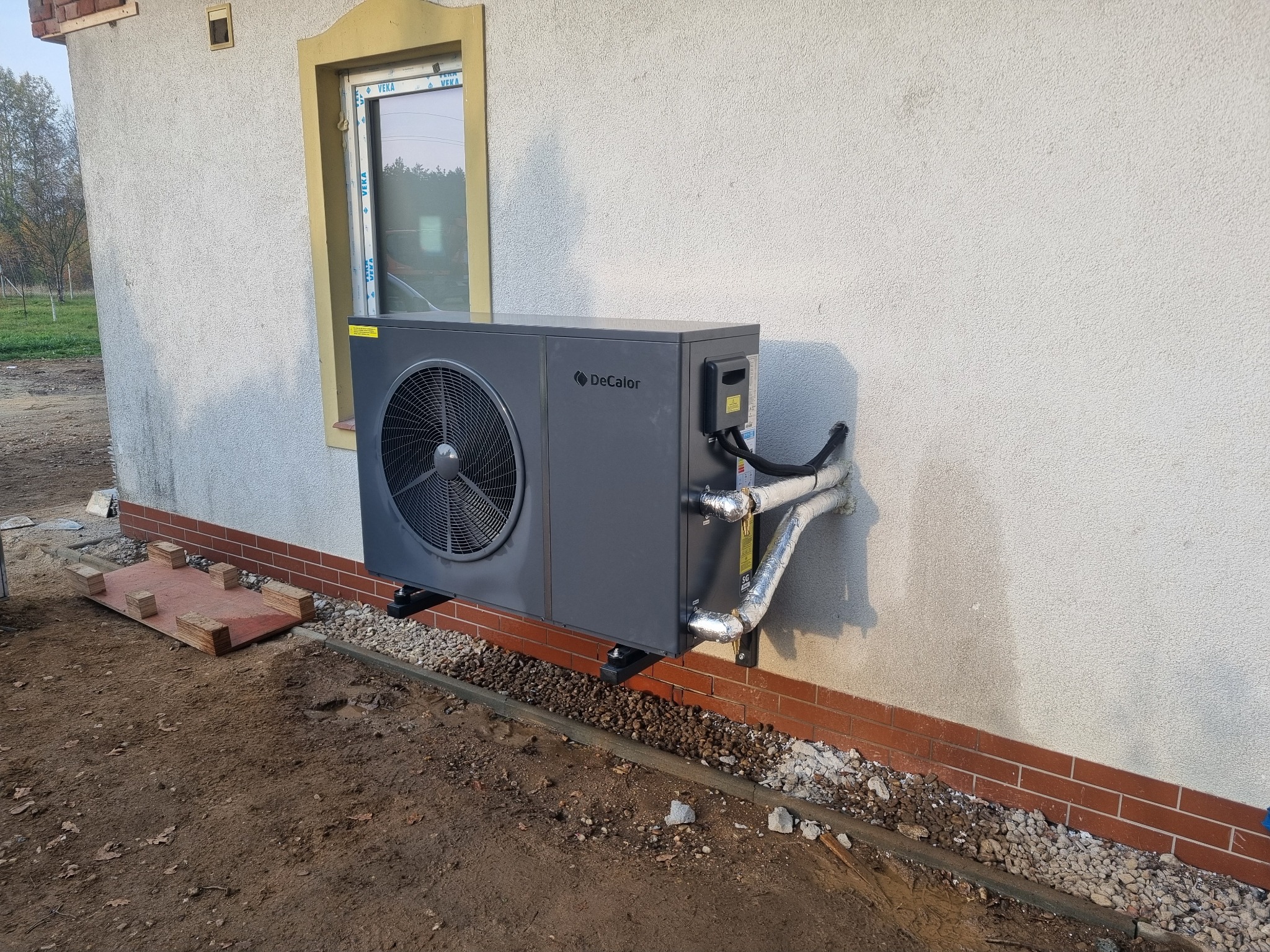 Szara pompa ciepła DeCalor zamontowana na ścianie budynku, widoczne rury w izolacji, teren wokół w trakcie prac wykończeniowych.