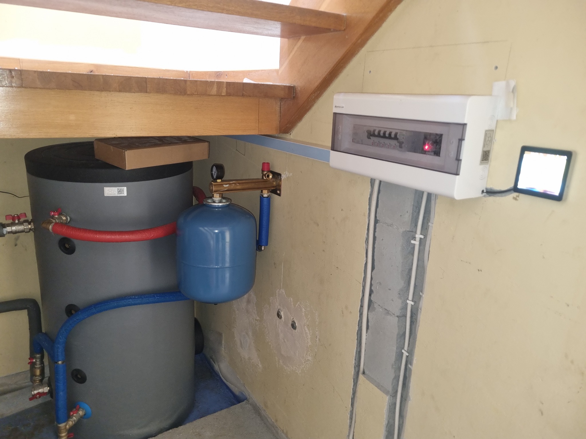 Szary zasobnik ciepłej wody z niebieskim naczyniem przeponowym, czerwonym i niebieskim orurowaniem, skrzynka elektryczna z panelem sterowania w tle, montaż w pomieszczeniu pod schodami.