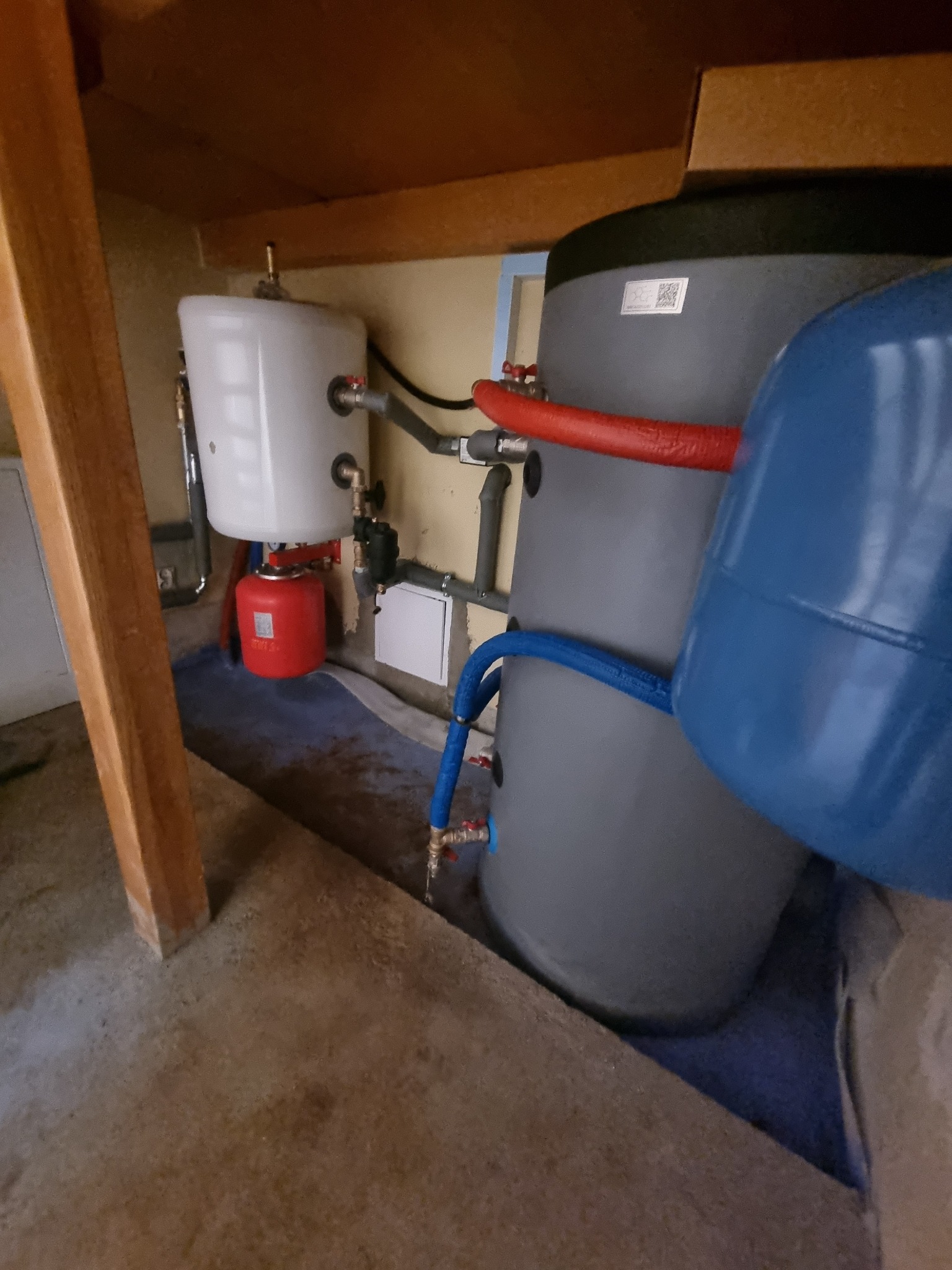 Instalacja dwóch zbiorników: biały mniejszy i szary większy, połączonych rurami w kolorach czerwonym i niebieskim, umieszczona pod drewnianą konstrukcją, prawdopodobnie w piwnicy.