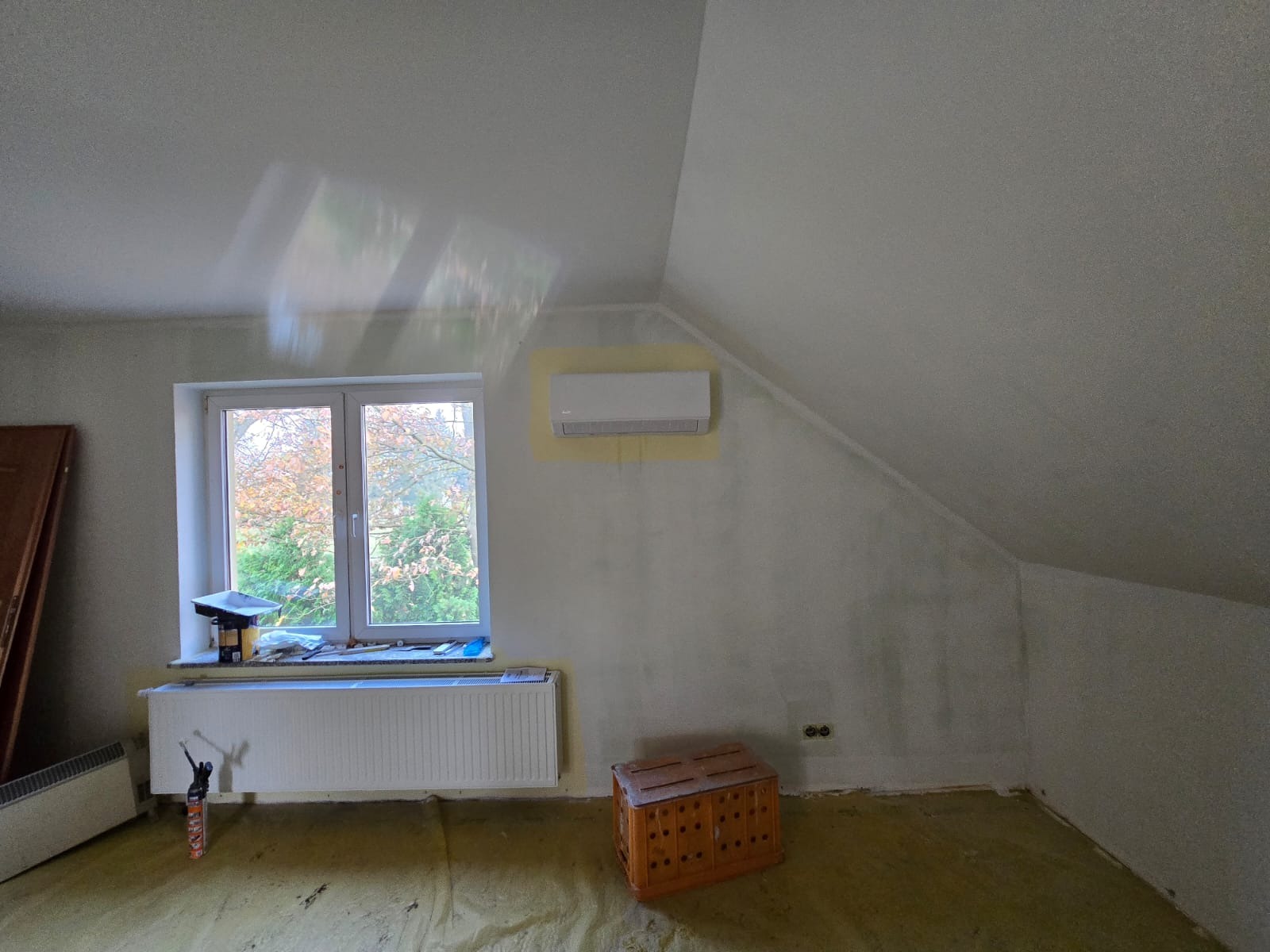 Wnętrze poddasza w trakcie remontu z zamontowaną jednostką wewnętrzną klimatyzacji na ścianie, widoczne okno z widokiem na drzewa, grzejnik i skrzynka.