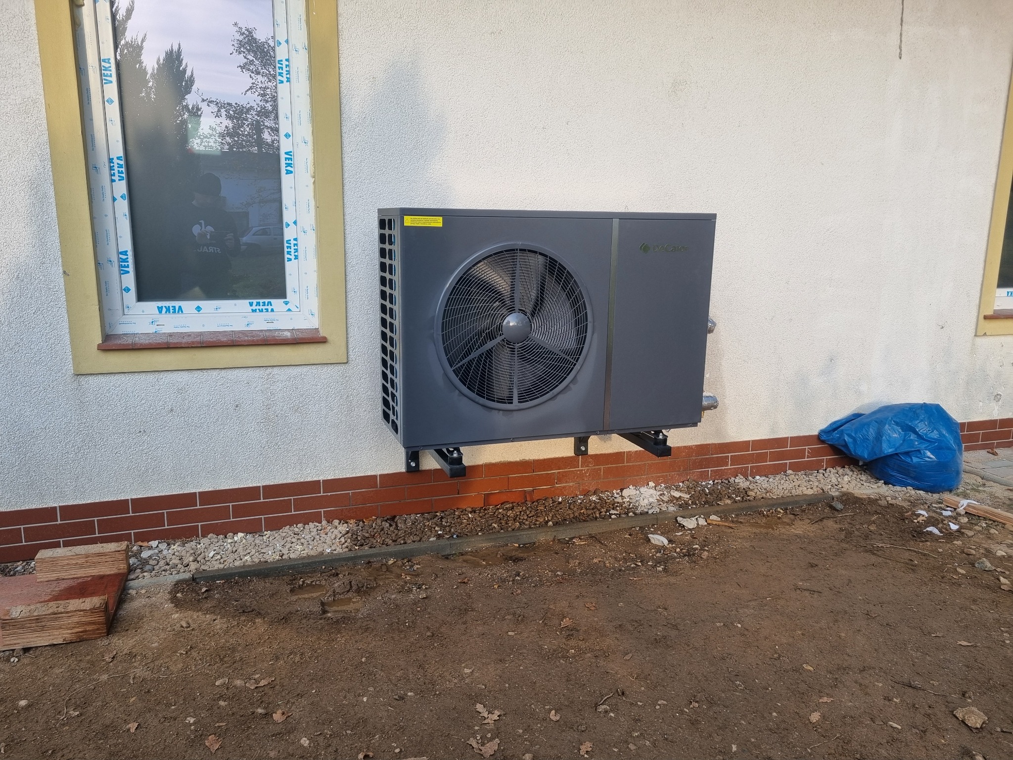 Szara pompa ciepła zamontowana na ścianie budynku z ceglaną podmurówką, widoczna z zewnątrz, z oknem w tle.