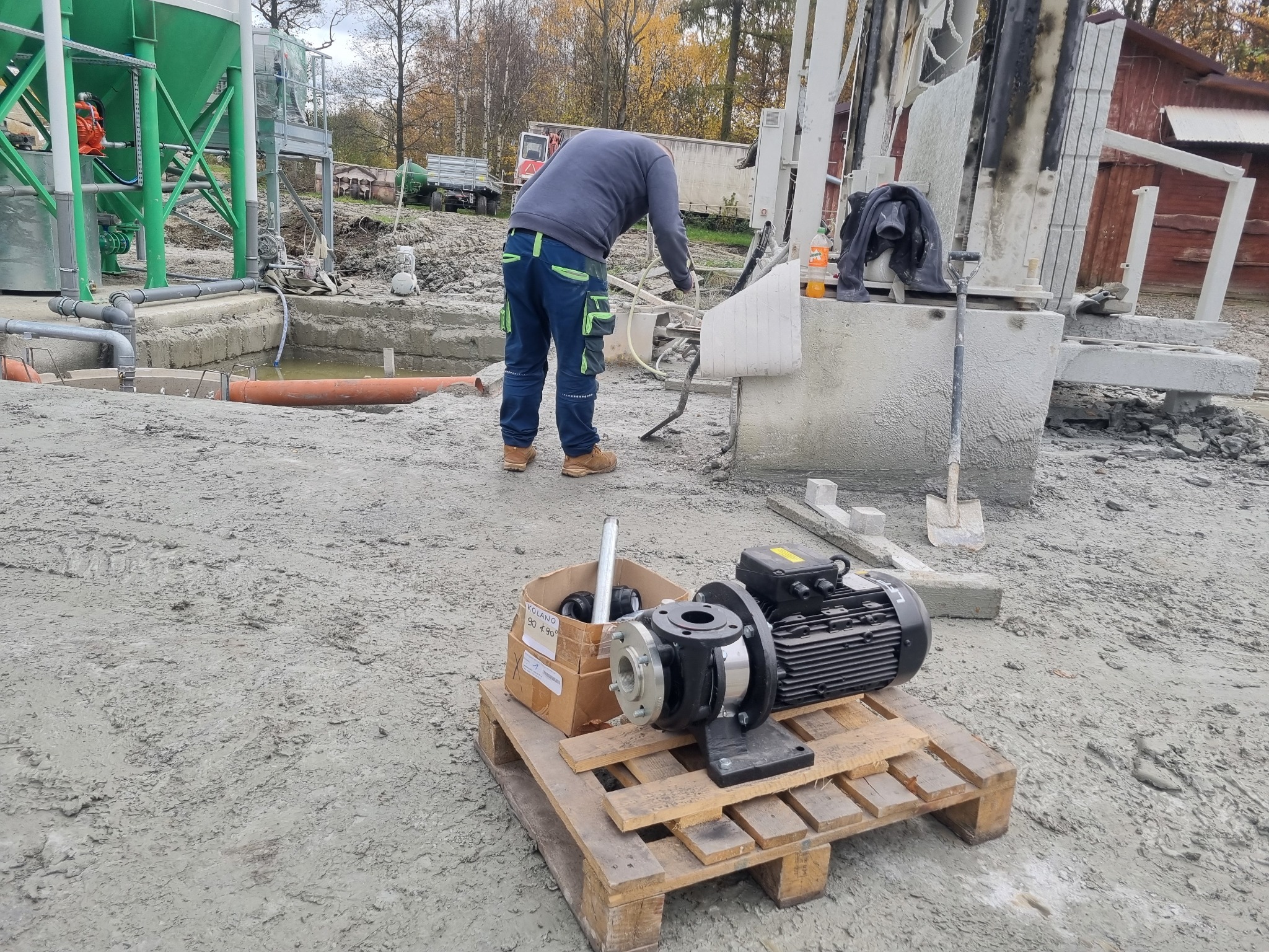 Montaż pompy ciepła: pracownik w ubraniu roboczym przy wiertnicy, pompa i akcesoria na palecie, zbiornik i instalacja w tle.