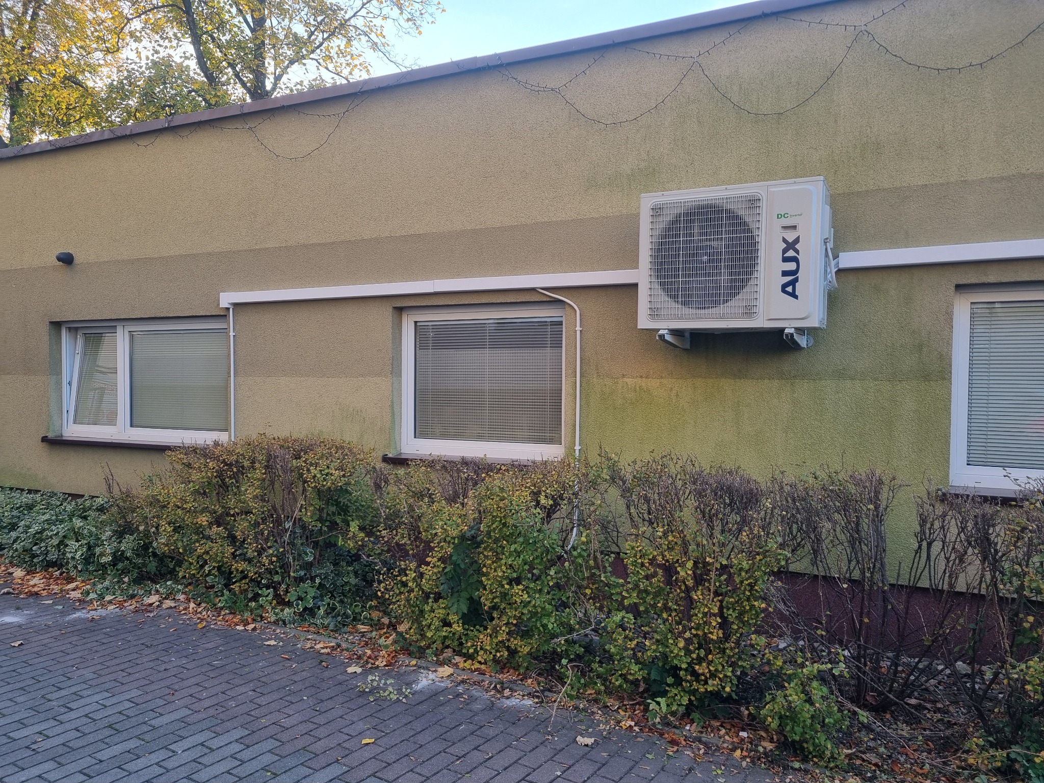 Zewnętrzna jednostka klimatyzacji marki AUX zamontowana na elewacji budynku w Gryfowie Śląskim, widoczna z chodnika, z krzewami u podstawy ściany.