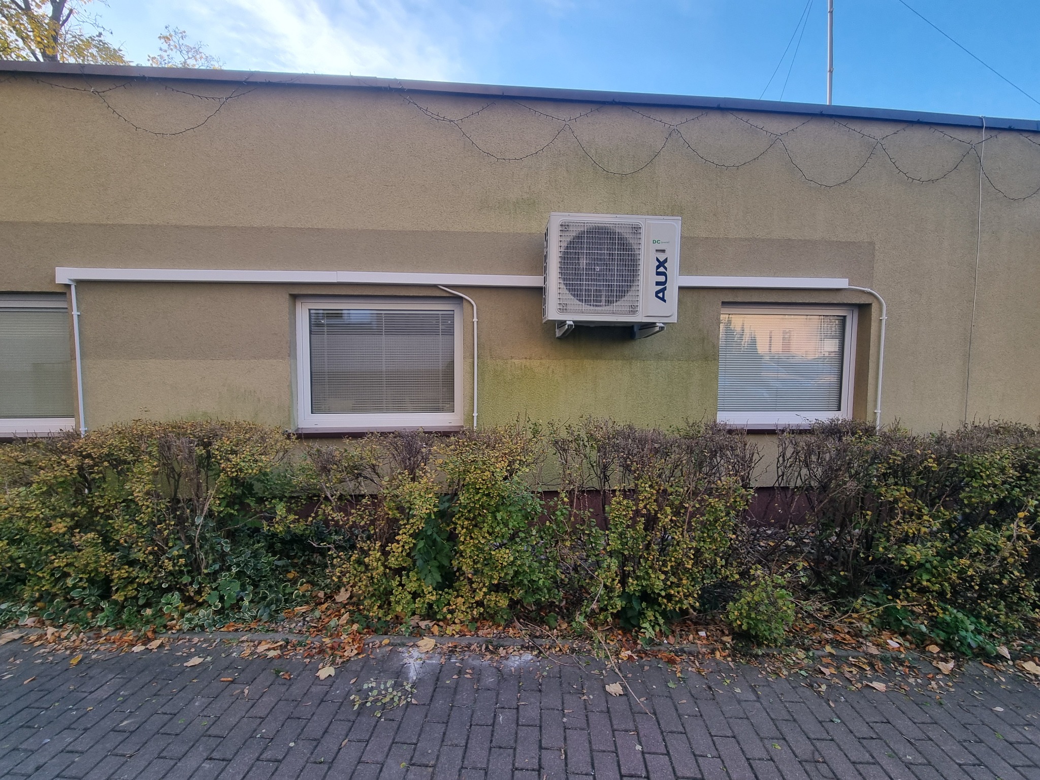 Biała jednostka zewnętrzna klimatyzacji marki AUX zamontowana na elewacji budynku, widoczna z chodnika, z krzewami u podstawy ściany.