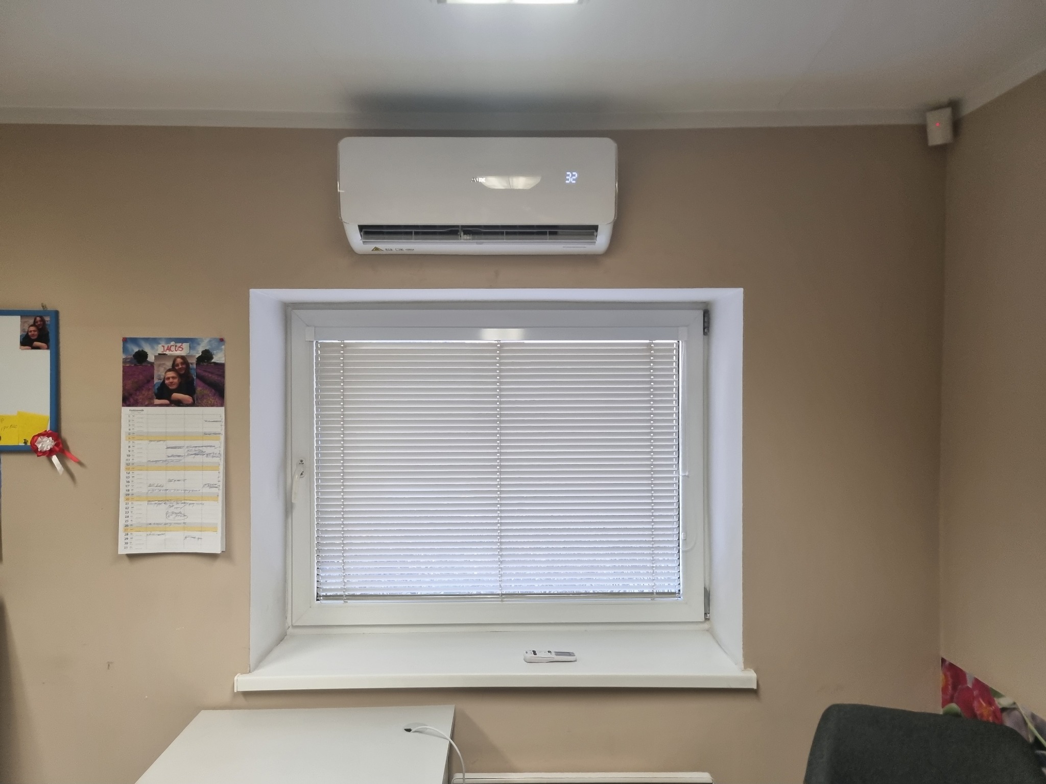 Biała klimatyzacja na ścianie nad oknem z żaluzjami w pomieszczeniu biurowym, widoczny kalendarz i fragment biurka.
