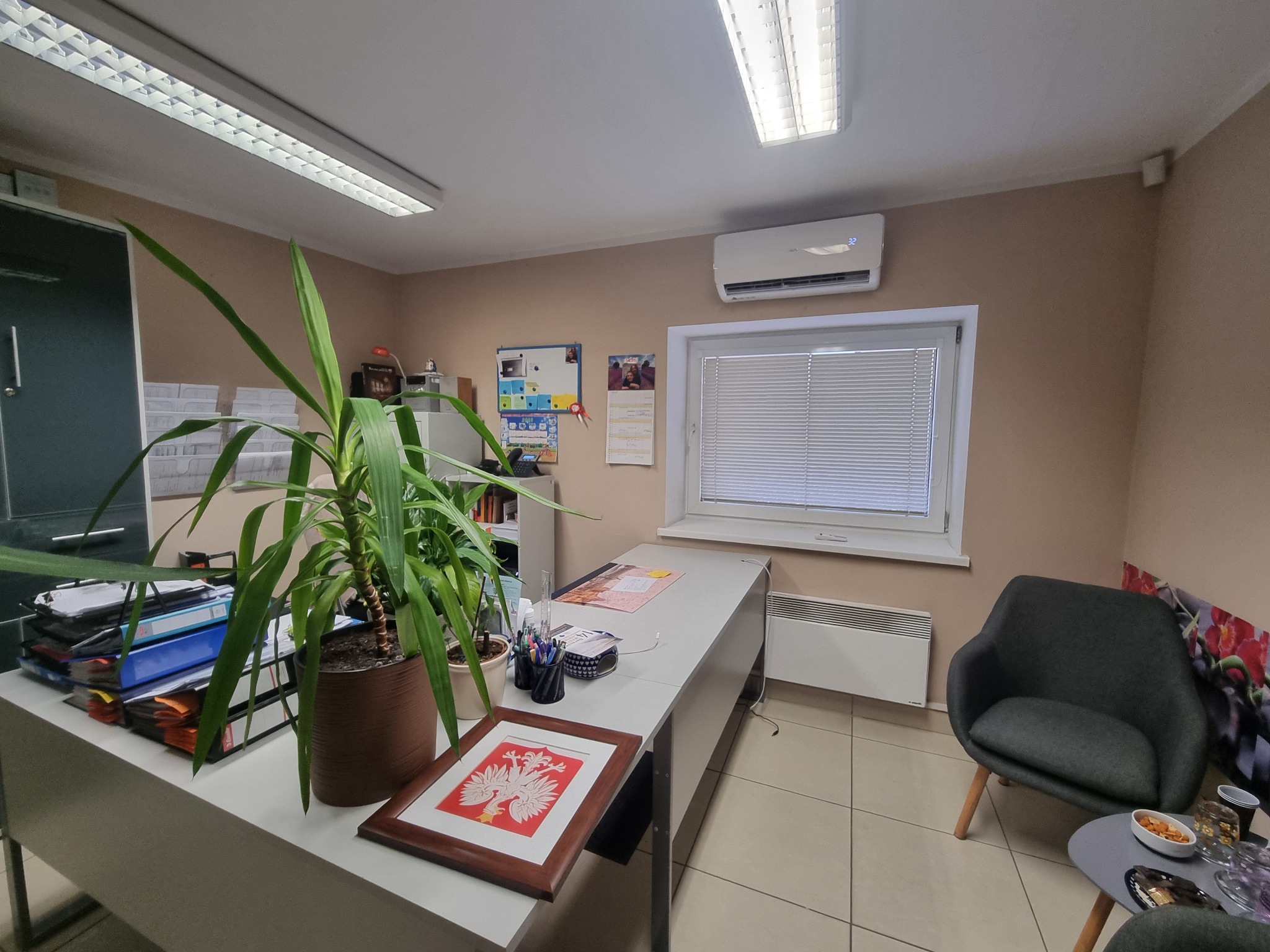 Wnętrze biura z klimatyzatorem, biurkiem, kwiatami doniczkowymi, krzesłem i godłem Polski w ramce.