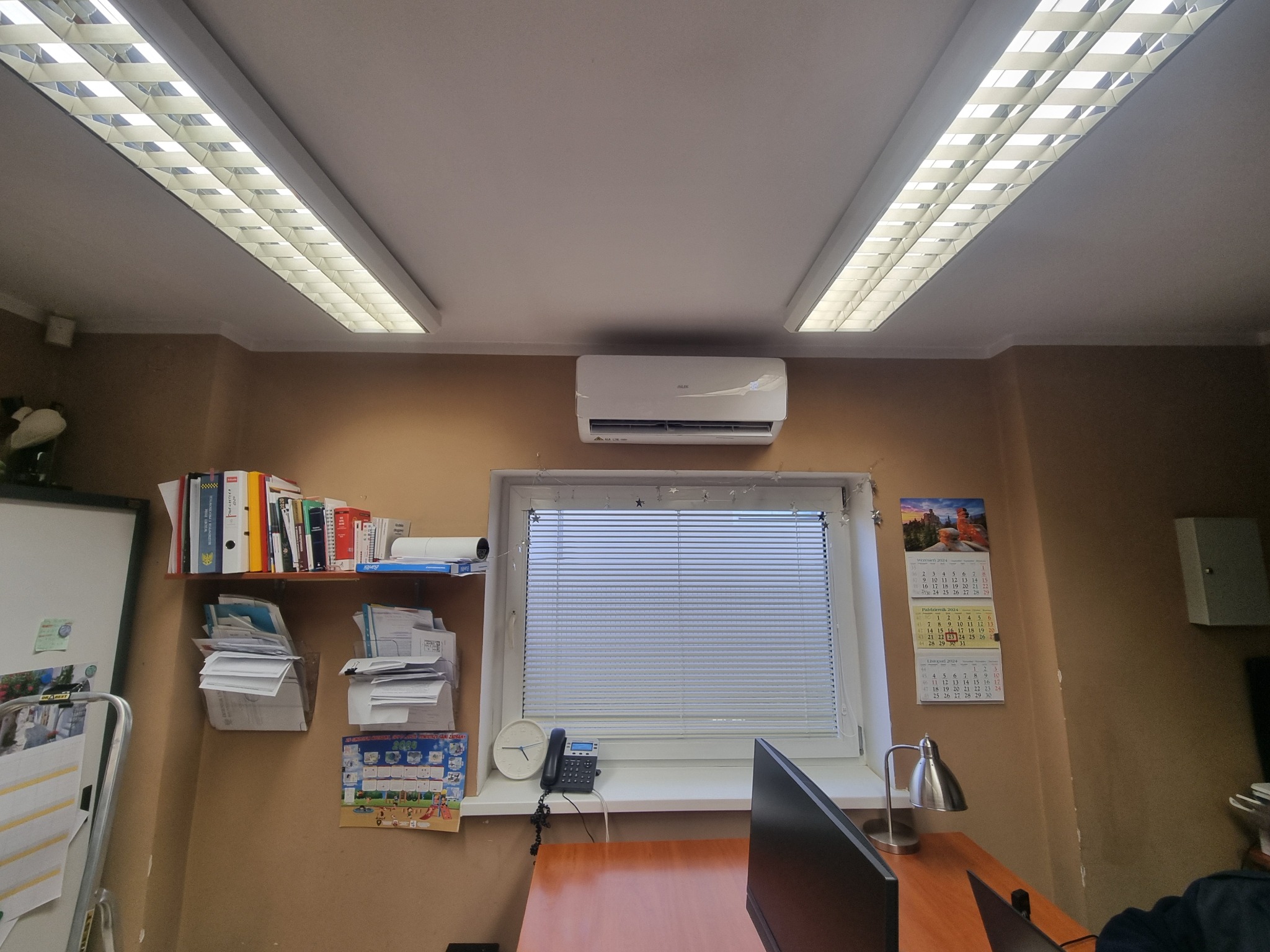 Wnętrze biura z klimatyzatorem zamontowanym nad oknem z żaluzjami, oświetlone podwójnym rzędem lamp sufitowych, z biurkiem, komputerem, półkami z dokumentami i kalendarzami na ścianie.