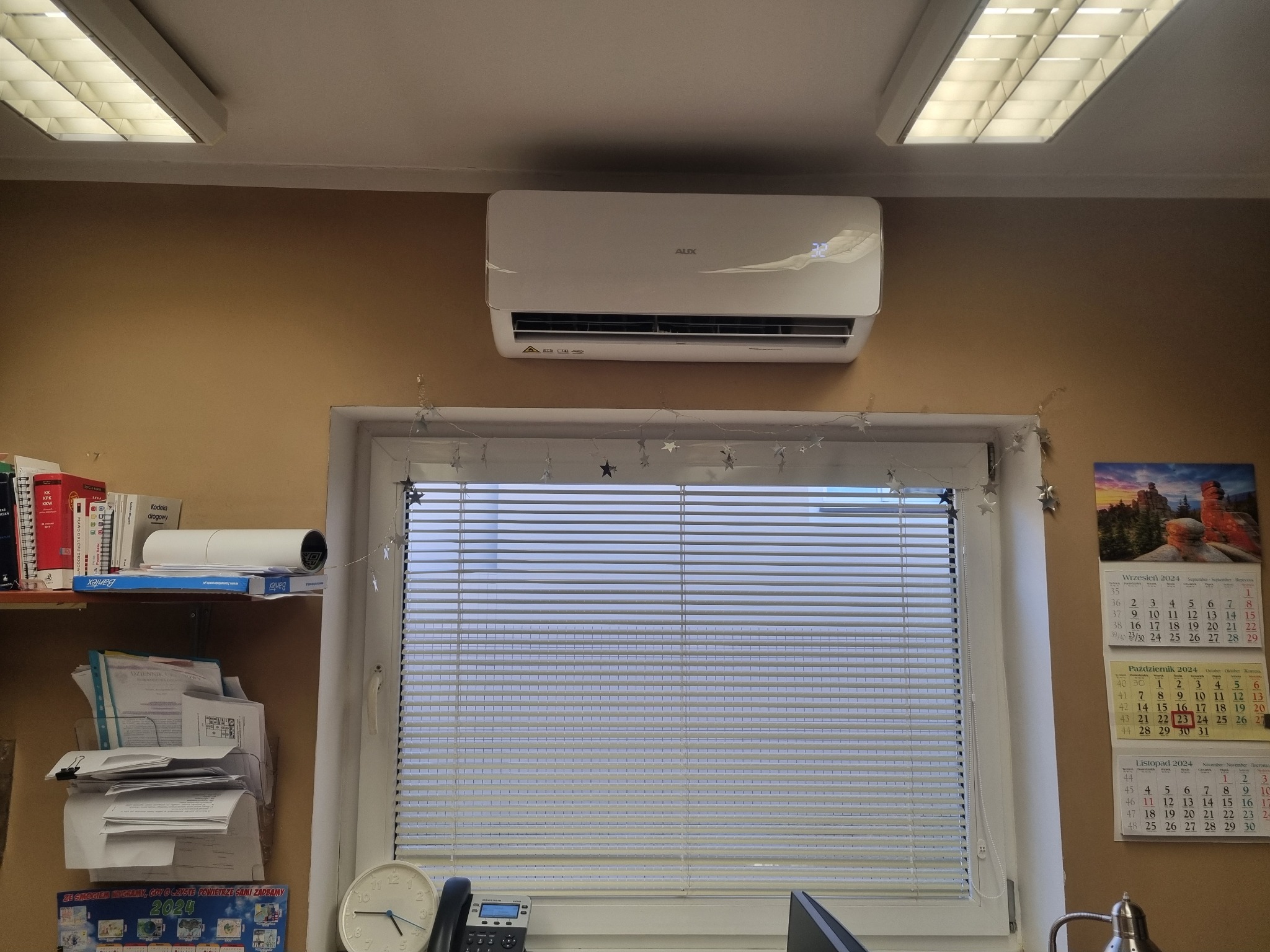 Klimatyzator marki AUX zamontowany na ścianie w pomieszczeniu biurowym, widok od dołu, z widocznymi żaluzjami w oknie i kalendarzem na ścianie.