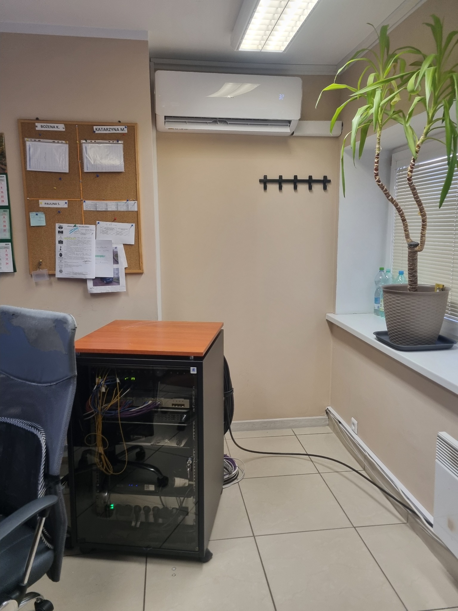 Wnętrze biura z klimatyzatorem zamontowanym na ścianie, szafą rack z widocznymi kablami, tablicą korkową z dokumentami i doniczką z rośliną na parapecie.