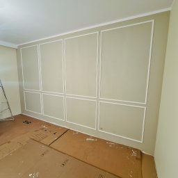 APARTMENTS REAL ESTATE SPÓŁKA Z OGRANICZONĄ ODPOWIEDZIALNOŚCIĄ - Wykończone wnętrze pokoju z beżowymi ścianami i białą sztukaterią w formie paneli; podłoga zabezpieczona kartonem, drabina aluminiowa w rogu.